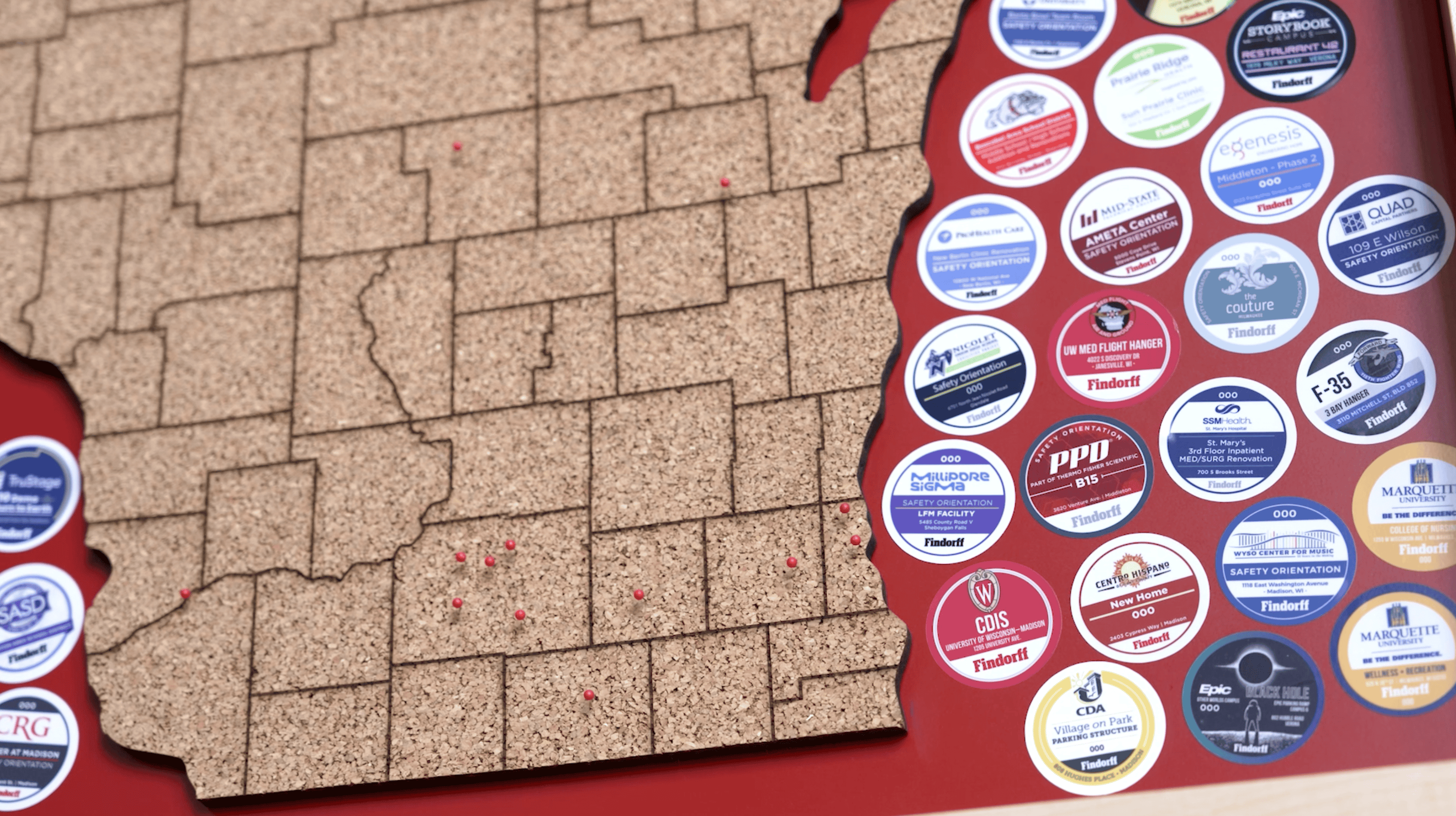 Close-up of a corkboard map of Wisconsin with red pins marking locations. Surrounding the map are colorful circular badges, each representing a different Findorff project or partnership.